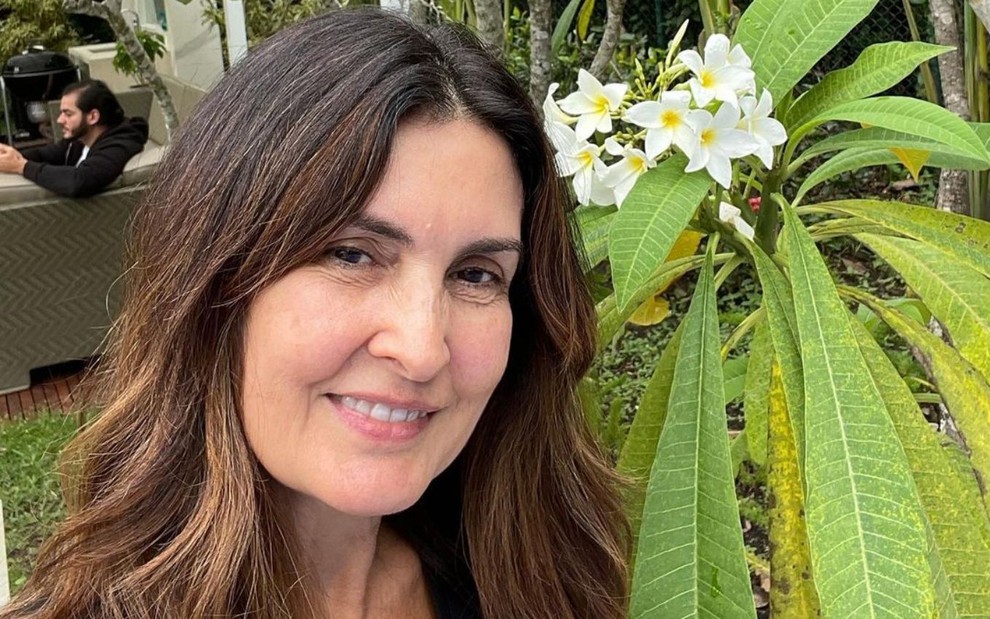 Jornalista Fátima Bernardes sorri ao lado de uma flor branca com folhas bem verdes