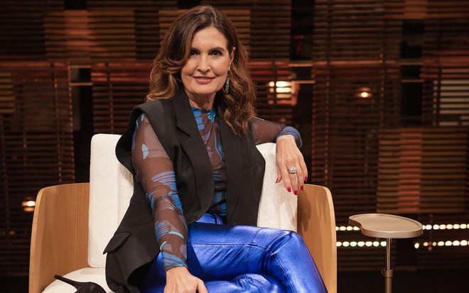 Com calça azul, colete preto e blusa estampada azul, Fátima Bernardes posa sentada em uma cadeira