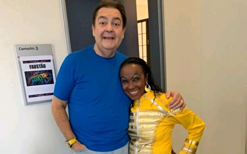 Mary Jackson abraçada a Fausto Silva em frente a camarim na Globo, ele de camiseta azul e ela de blusa amarela; ambos sorrindo