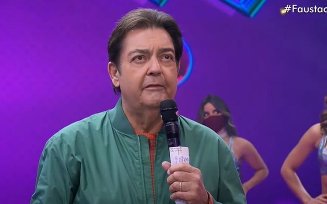 Fausto Silva usa um agasalho verde e segura o microfone enquanto comanda o seu Faustão na Band