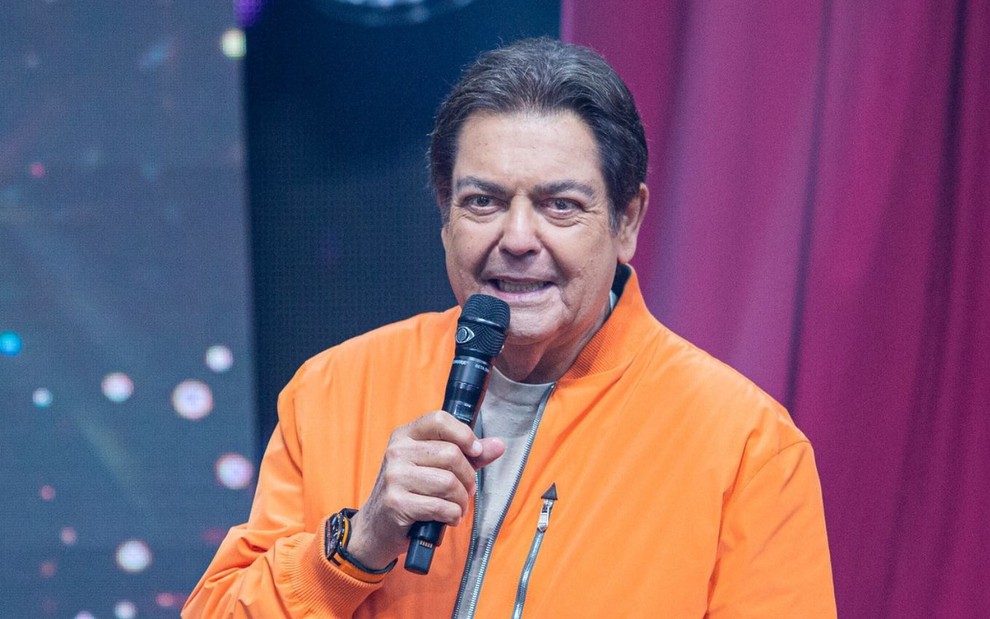 De jaqueta laranja, Faustão segura microfone com a logomarca da Band