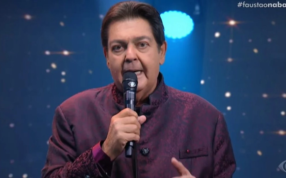 O apresentador Fausto Silva de terno roxo, microfone na mão, expressão séria, em estúdio de seu programa na Band