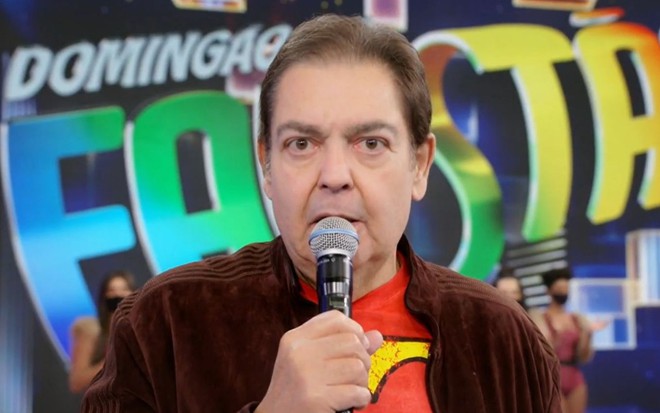 Com jaqueta marrom e camiseta vermelha, Fausto Silva tem expressão de choque ao olhar para a câmera