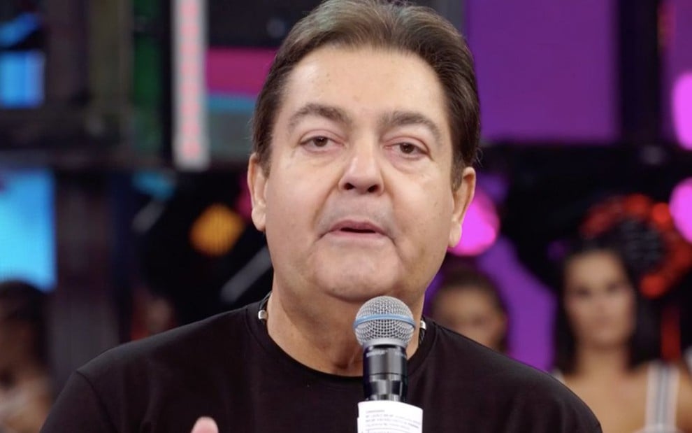 Fausto Silva no palco do Domingão, de camiseta preta, segurando microfone