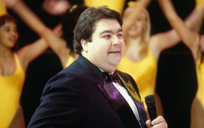 Fausto Silva usa terno e está com microfone na mão; atrás aparecem bailarinas de collant amarelo