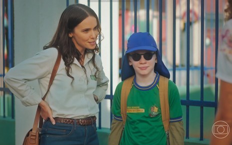 Em cena de Terra e Paixão, Débora Falabella está ao lado de Felipe Melquiades na porta da escola; ele usa óculos escuros e boné azul