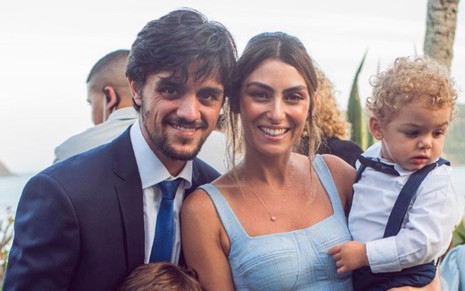 Felipe Simas, Mariana Uhlmann e seu filho mais novo, Vicente, posam sorrindo para foto. Felipe veste um terno preto com gravata azul e camisa branca. Mariana veste um vestido azul. O filho mais novo está de suspensórios e gravata borboleta.