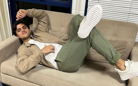 Felipe Simas está deitado no sofá de um camarim da Globo: ele usa camiseta branca, um blusão caramelo por cima, calça verde e tênis branco