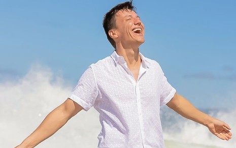 Felipe Suhre na foto de capa de seu livro, com camisa branca de manga curta, braços abertos, sorrindo em paisagem externa, sob o sol