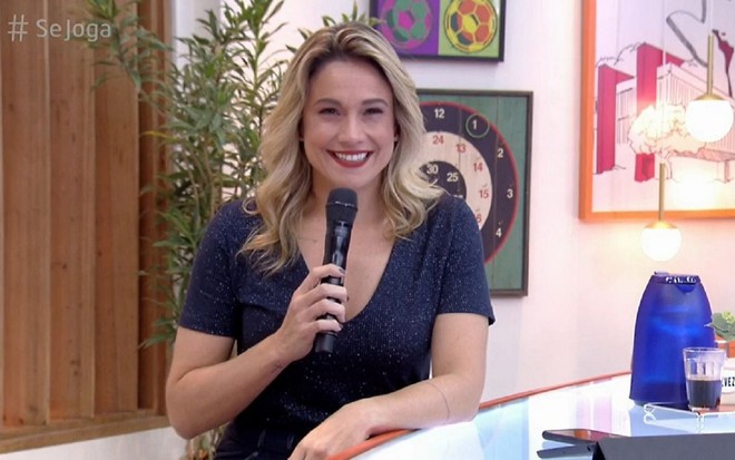 Fernanda Gentil usa uma camisa azul e está com um microfone preto na mão. Ela está sentada e usa também um batom vermelho no cenário do Se Joga de sábado.