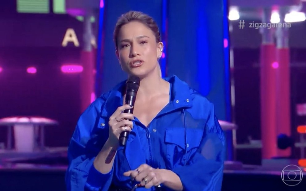 Fernanda Gentil usa uma camisa azul e está com um microfone preto na mão. Ela está no cenário do Zig Zag Arena.