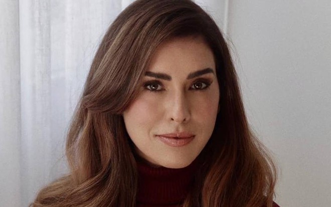 Fernanda olha para frente, está com o cabelo solto e está na frente de uma parede branca
