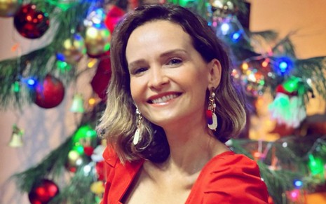 A atriz Fernanda Rodrigues olha para a câmera, sorri, de blusa vermelha, em frente a árvore de Natal