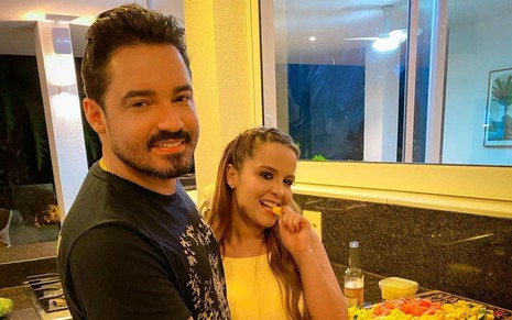 Fernando Zor e Maiara estão em uma cozinha e posam para uma foto no Instagram. Fernando está sorrindo de camiseta preta, e Maiara veste um body amarelo e posa comendo um pedaço de fruta