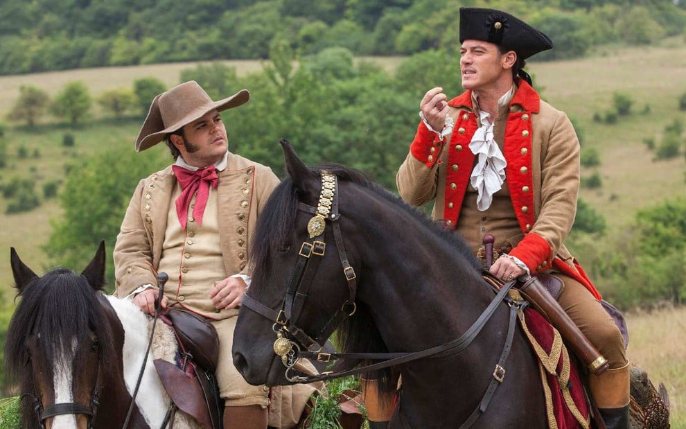 LeFou (Josh Gad) na esquerda e Gaston (Luke Evans) na direita conversam enquanto andam à cavalo em cenário sem ninguém ao fundo em cena do filme A Bela e a Fera (2017)