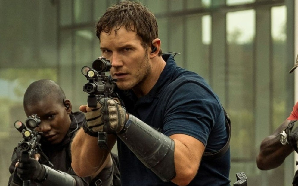 Dan (Chris Pratt) no lado direito de camiseta azul segurando uma arma com as duas mãos e de olho na mira e uma mulher no lado esquerdo com a mesma arma olhando para frente em cena de A Guerra do Amanhã