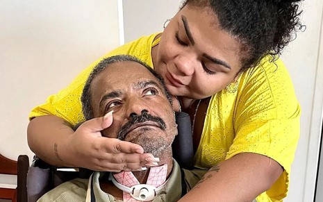 De blusa amarela, Flora Cruz abraça o rosto do pai, Arlindo Cruz, que está olhando para ela