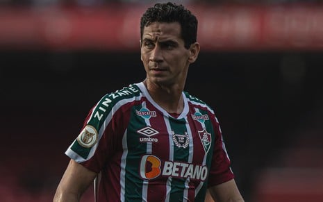 Ganso, do Fluminense, caminha em campo e veste uniforme listrado em grená, verde e branco