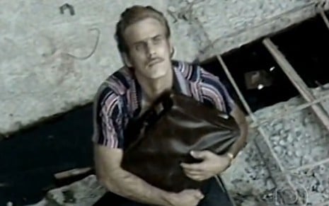 Francisco Cuoco segura uma mala e olha para cima em cena como Carlão da novela Pecado Capital (1975)