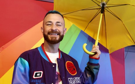 Fred Bruno us um casaco roxo e azul e uma camisa branca; ele sorri para a câmera enquanto segura um guarda-chuva cenográfico. Ao fundo, é possível ver um backdrop com as sete cores do arco-íris.