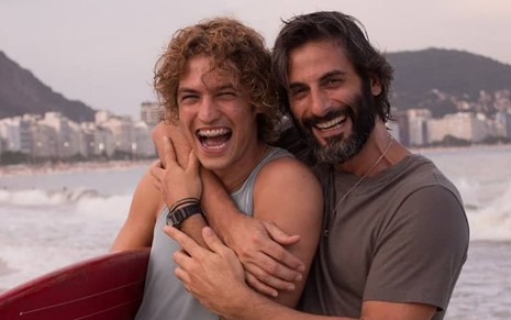 Gabriel Leone e Flavio Tolezani estão em frente ao mar; Gabriel segura uma prancha vermelha e veste regata cinza; Flavio está ao lado de Gabriel e veste camiseta cinza; os dois estão sorrindo