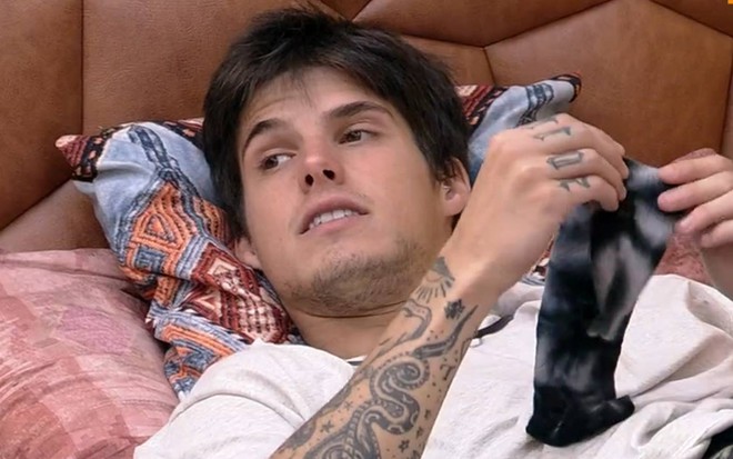 Foto de Gabriel; ele está deitado na cama e veste camiseta branca