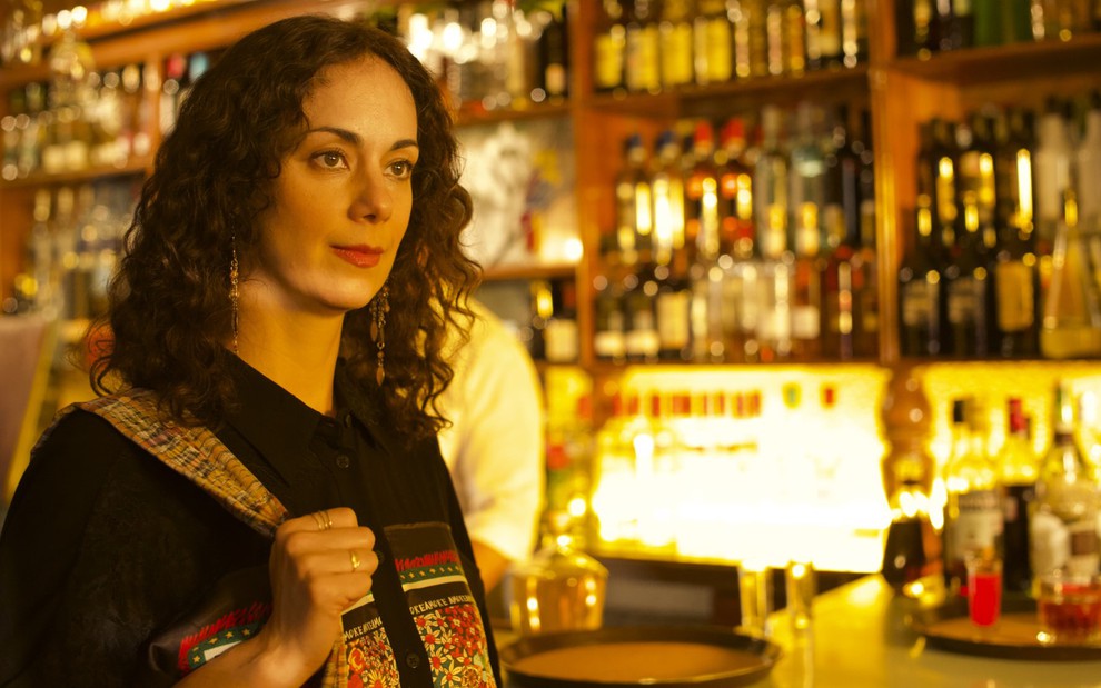Gabriela de la Garza segurando uma bolsa, vestindo uma blusa preta, ao lado de um balcão de um bar