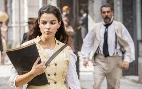 Pilar (Gabriela Medvedovski) caminha pela rua; Tonico (Alexandre Nero) aparece atrás dela em cena de Nos Tempos do Imperador