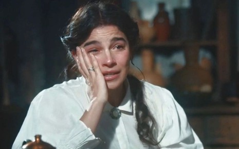 Gabriela Medvedovski coloca a mão direita no rosto e limpa lágrimas em cena como Pilar na novela Nos Tempos do Imperador