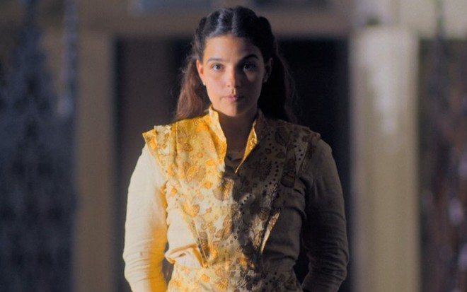 Gabriela Medvedovski usa um vestido amarelo e está com expressão séria em cena como Pilar de Nos Tempos do Imperador