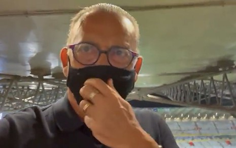 Galvão Bueno leva a mão à máscara, com os olhos cheios d'água; ele está em um estádio de futebol