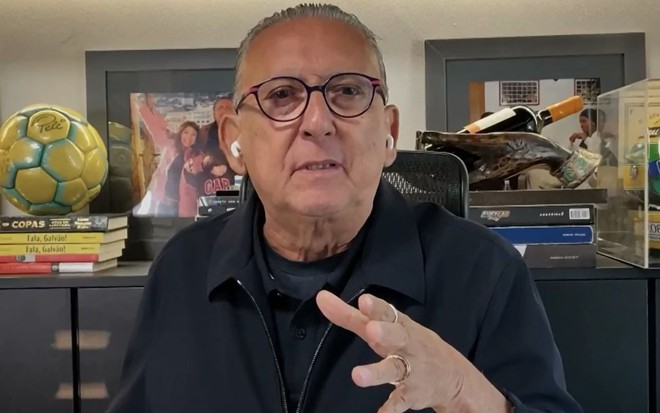 Galvão Bueno com camisa preta, óculos, olhando para a câmera; ao fundo, bola autografada por Pelé e capacete de Ayrton Senna