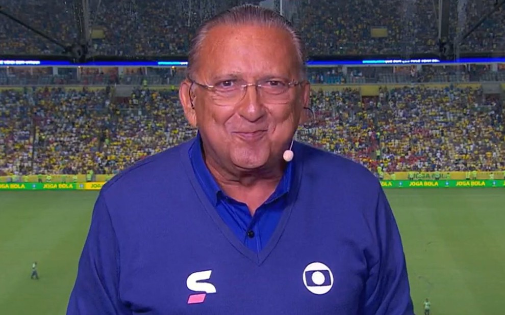 Galvão Bueno com uma blusa azul e calça marrom, em um fundo cinza, sorrindo para a câmera em uma transmissão da Globo