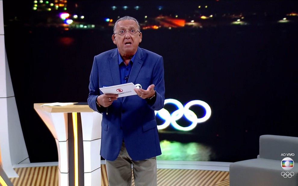 Galvão Bueno no espaço virtual da Globo montado para os Jogos Olímpicos nos Estúdios Globo. Todos usam ternos azuis com o logotipo da Globo e calça marrom.