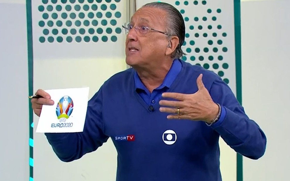 No estúdio esportivo da Globo, Galvão Bueno segura uma ficha da Euro 2020; ele está de uniforme azul