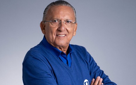 Galvão Bueno com uma blusa azul e calça marrom, em um fundo cinza, sorrindo para a câmera em uma foto divulgada pela Globo