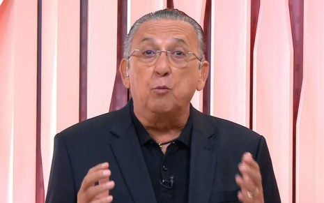 Galvão Bueno, vestido com terno preto e camisa preta, em um fundo de estúdio rosa, falando sobre futebol nos estúdios da Globo