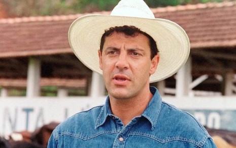 Gerson Brenner em cena de Corpo Dourado (1998) com camisa jeans, chapéu branco e expressão séria