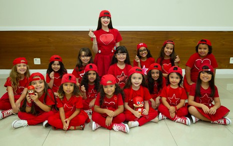 Vestida com uma blusa vermelha com coração e boné da mesma cor, Gi Alparone fotografa ao lado de 15 meninas vestidas da mesma forma