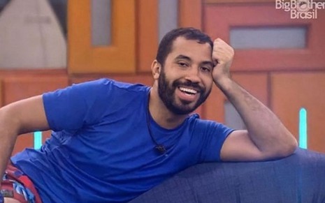 Gil do Vigor veste uma camiseta azul enquanto descansa em um sofá no jardim da casa onde é gravado o Big Brother Brasil nos estúdios Globo