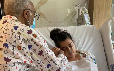Gilberto Gil durante visita à filha Preta Gil no hospital, ela está deitada em cama, com sonda no nariz, segurando a mão do pai, que está de pé