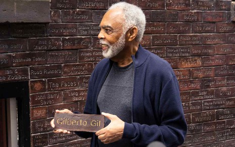 Gilberto Gil sorrindo, segurando placa com seu nome em frente a muro cheio de placas com nomes de artistas da música