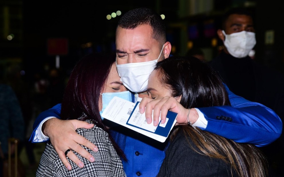 Gil do Vigor chorando enquanto abraça a mãe (à esq.) e a irmã no aeroporto de Guarulhos, em São Paulo