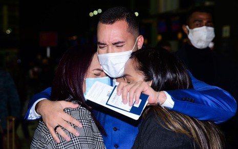 Gil do Vigor chorando enquanto abraça a mãe (à esq.) e a irmã no aeroporto de Guarulhos, em São Paulo