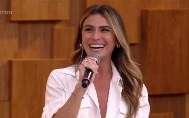 Giovanna Antonelli no estúdio do Encontro com Fátima Bernardes vestindo uma camisa branca e segurando o microfone com a mão direita. A atriz está sorrindo.