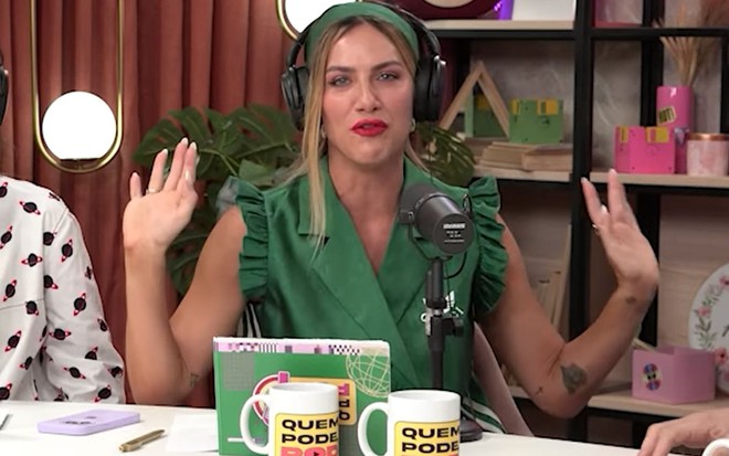 A atriz e apresentadora Giovanna Ewbank durante apresentação do podcast Quem Pode Pod; ela está sentada, olhando para a câmera com um fone de ouvido e falando ao microfone com as duas mãos para cima