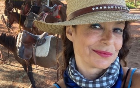 A atriz Giovanna Gold com chapéu de cowboy, lenço azul no pescoço, faz selfie em frente a cavalos