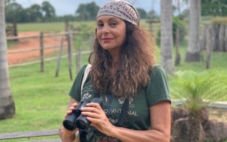 A atriz Giovanna Gold em cenário de campo, com binóculo nas mãos, bandana na cabeça, olha para a câmera
