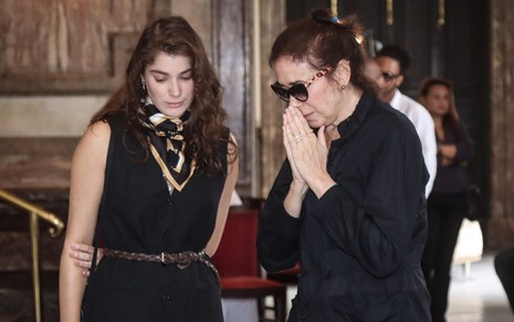 Giulia Bertolli ao lado de Lilia Cabral, que junta as mãos em oração; ambas vestem preto
