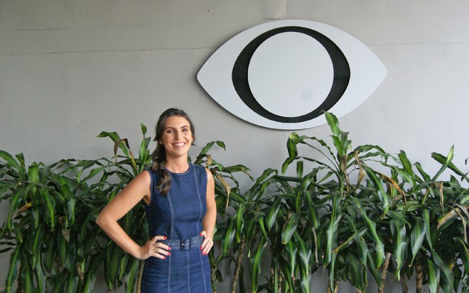Glenda Kozlowski em um vestido jeans, na frente da sede da Band, com plantas e o logotipo da emissora atrás